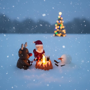 Santa, rendier en sneeuwpop zitten bij een gloeiend kampvuur in de sneeuw terwijl kerstlampjes twinkelen op de achtergrond. Winterse sfeer met humoristische twist — unieke handgemaakte kerstdecoratie voor binnen of onder de boom.