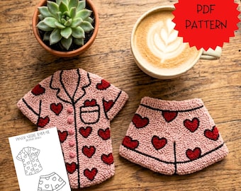 May include: A punch needle pyjama set featuring a pink short-sleeve button-down shirt and matching shorts with a red heart pattern. A white card displays the line art template labelled PDF PATTERN, alongside a succulent and a coffee mug.