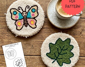 May include: Two cream-coloured, round coasters. One has a colourful butterfly design, the other a green leaf. A cup of coffee sits on a saucer in the background. A paper with a butterfly and leaf template is also present. The coasters are likely 10 cm in diameter.