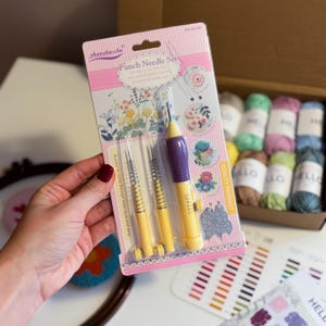 May include: A set of four punch needles for embroidery, packaged in a pink and white plastic sleeve. The needles are yellow and purple with a spring mechanism. The packaging includes a colour chart of embroidery floss.