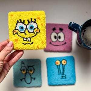 May include: Four square coasters featuring characters from the cartoon Spongebob Squarepants. The coasters are made of a soft, fuzzy material and are yellow, pink, blue, and light blue. The characters are Spongebob, Patrick Star, Squidward Tentacles, and Plankton.