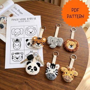 Puede incluir: Una colección de llaveros hechos a mano con temática de animales, incluyendo un tigre, un elefante, un león, un panda, una cebra y una jirafa. Los llaveros están hechos con tela texturizada y unidos a una correa beige con un anillo de metal. Una "Punch Needle Template" es visible.