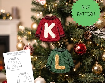 Op de afbeelding: Een punch needle PDF-patroon met twee ornamenten in de vorm van een trui met de letters K en L. De rode trui heeft een witte K en de groene trui heeft een gele L. Een wit papieren sjabloon toont de contouren van de truien met de letters.