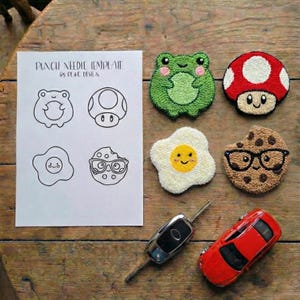 May include: A selection of colourful, handmade punch needle designs, including a green frog, red mushroom, fried egg, and cookie, next to a car key and a red toy car. A paper with punch needle template designs is also present.