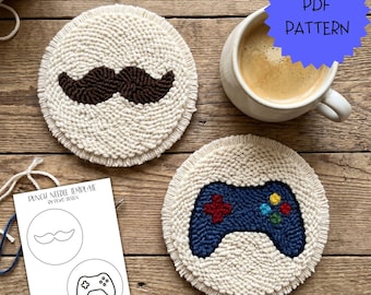 May include: Two circular punch needle coasters with cream-coloured yarn backgrounds. One features a dark brown moustache, the other a blue video game controller with red, yellow, green, and blue buttons. Includes a paper template labelled PUNCH NEEDLE TEMPLATE.