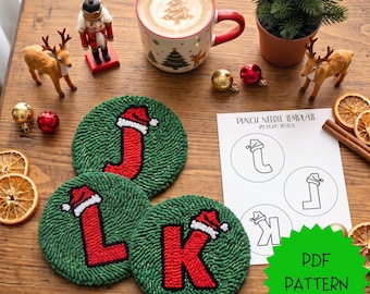 Op de afbeelding: Drie ronde punch needle onderzetters in groen met rode letters J, L en K voorzien van kerstmutsen met witte randen. Een wit vel papier toont een punch needle sjabloon met de letters J, L en K, en een felgroene sterfiguur met de tekst PDF PATTERN.