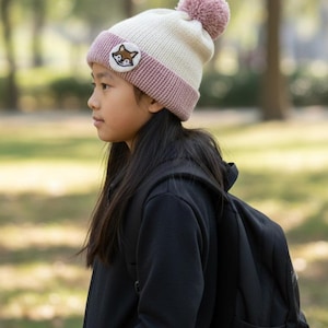 May include: A knitted winter hat with a fox patch and a pom-pom. The hat features a cream-colored crown, a mauve band, and a matching pom-pom. A circular patch with a fox face is sewn on the front. The hat is designed for cold weather.