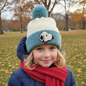 May include: A child wearing a knit hat with a blue brim and a Snoopy design, a matching blue pom-pom, and a red scarf. The hat is cream-colored and the child is wearing a navy blue jacket. The background is a park with fall foliage.