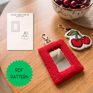 Puede incluir: Un espejo rojo, rectangular y texturizado con un llavero dorado. También hay un llavero en forma de cereza blanca y roja. Un cuenco de cerezas y una plantilla de aguja de perforación con un diseño de cereza están sobre una superficie de madera. El texto "PDF PATTERN" es visible.