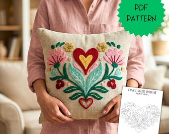 May include: A square beige throw pillow featuring a central punch needle embroidery design of a red and yellow heart surrounded by pink flowers, green leaves, and small red and yellow accents. A white paper template shows the line art pattern.