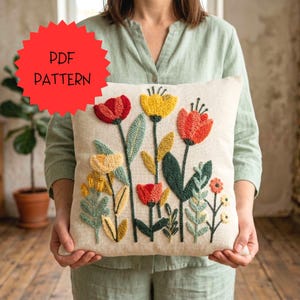 Pode incluir: Uma almofada quadrada bege com um design floral colorido. As flores são vermelhas, amarelas e laranja, com folhas verdes e amarelas. A almofada tem uma aparência texturizada. Uma forma de estrela vermelha com as palavras "PDF PATTERN" está no canto superior esquerdo.