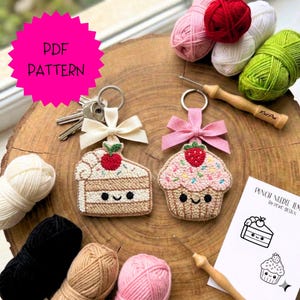 Pode incluir: Dois chaveiros feitos à mão, um em forma de fatia de bolo e outro de cupcake, ambos com rostos sorridentes e laços. Os chaveiros são feitos com fio e têm um anel de metal. A imagem também inclui novelos de lã e um sinal de "PDF PATTERN".