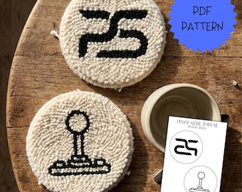 May include: Two round punch needle coasters with cream-coloured yarn and black yarn designs, one featuring a stylised logo and the other a joystick icon. A white paper sheet displays the text PUNCH NEEDLE TEMPLATE BY PCWD DESIGN with matching line art.