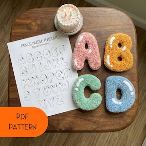 May include: A wooden table displays a punch needle alphabet template with the letters A, B, C, and D in pink, orange, green, and blue. A small cake with candles sits at the top. The text "PDF PATTERN" is visible.