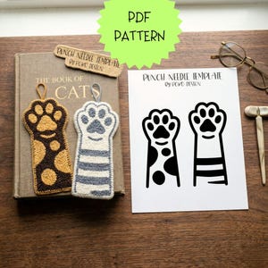 Op de afbeelding: Op een houten tafel ligt een boek met de titel "The Book of Cats" met twee kattenpoot-bladwijzers en een bord "PDF PATTERN". Er ligt ook een papier met een kattenpoot sjabloon. De bladwijzers zijn bruin en grijs.
