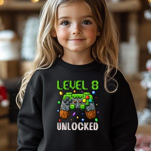 Op de afbeelding: Zwart sweatshirt met een groene en witte gepixelde videogamecontroller afbeelding. De tekst "Level 8 Unlocked" is onder de controller gedrukt.