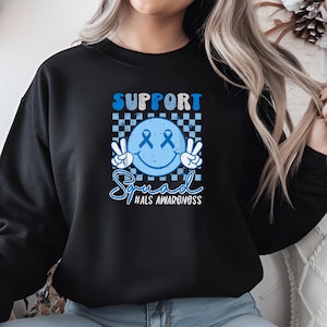 May include: Black sweatshirt with a blue smiley face with peace signs and blue ribbons for eyes. The text "SUPPORT Squad #ALS AWARENESS" is printed below the smiley face.