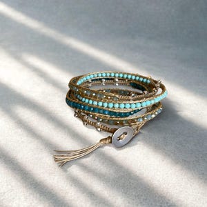 May include: Wrap bracelet featuring multiple strands of turquoise and silver beads, woven with a tan cord. The bracelet wraps around the wrist multiple times and fastens with a button closure and a tassel.