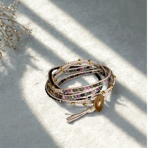 May include: Wrap bracelet featuring a combination of black, beige, and multi-colored beads. The bracelet is adorned with gold-colored accents and a button closure. The design includes a tassel detail, adding a touch of bohemian flair to this jewelry piece.