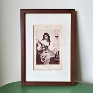 May include: A framed sepia-toned print of a woman seated, working with a spinning wheel. The print is in a dark brown wooden frame with a white mat. The woman is wearing a white blouse and striped skirt. The print is titled "Perrette, la fileuse."