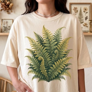 May include: An ivory t-shirt featuring a large, detailed graphic print of a lush green fern plant centered on the front. The shirt has a classic crew neckline, short sleeves, and a relaxed fit, showcasing botanical artwork with shades of green.