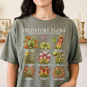 May include: A moss green t-shirt featuring a graphic print titled PREDATORY FLORA A NATURAL HISTORY OF LIVING TRAPS. The design displays nine detailed botanical illustrations of carnivorous plants with their common and scientific names.