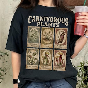 May include: Black t-shirt featuring the text "CARNIVOROUS PLANTS" above six postage stamp-style illustrations of various carnivorous plants. The shirt is a dark color and has a relaxed fit. The text "Black" is in a blue oval in the upper left corner.