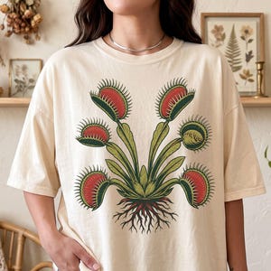 Könnte beinhalten: Ein kurzärmeliges T-Shirt in Elfenbein mit einer zentralen grafischen Illustration einer Venusfliegenfalle. Die Pflanze hat grüne Stängel und Blätter mit rot gefärbten, stacheligen Fallen sowie sichtbare braune Wurzeln an der Basis.