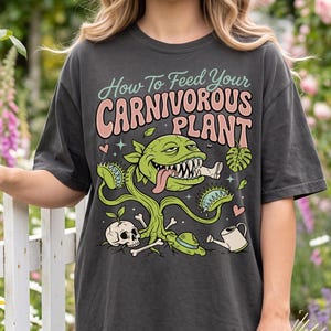 May include: A charcoal grey t-shirt featuring a graphic print with the text "How To Feed Your Carnivorous Plant" in pink and green. The design includes a green Venus flytrap, a human bone, a skull, a watering can, and small pink hearts.
