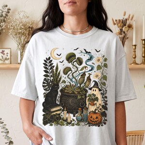 May include: A white t-shirt featuring a graphic print of a black cat sitting on books, a cauldron with carnivorous plants, a ghost wearing a witch hat, a jack-o'-lantern, candles, potion bottles, a crescent moon, and bats in a dark forest setting.