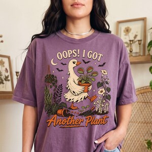 May include: A berry-colored t-shirt featuring a graphic of a white goose holding a potted Venus flytrap. The text reads OOPS! I GOT Another Plant. The design includes various potted plants, small bats, and a trowel on a dark soil patch.