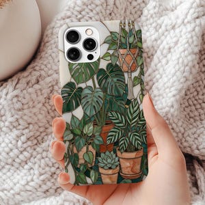 May include: A white phone case featuring a botanical illustration of various potted plants in shades of green and brown. The case includes a hanging plant, monstera leaves, and other greenery. The phone case is held in a person's hand.