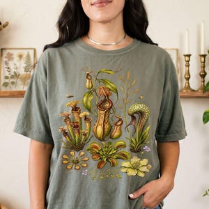 May include: A moss green short-sleeve t-shirt featuring a detailed botanical illustration of various carnivorous plants, including pitcher plants, a Venus flytrap, and sundews, rendered in shades of green, yellow, and brown across the chest.