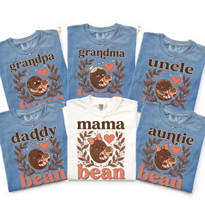 May include: Five blue and one white short-sleeve t-shirts featuring a graphic of a coffee bean character holding a baby. Each shirt displays text including grandpa, grandma, uncle, daddy, mama, or auntie bean, surrounded by leaves and hearts.