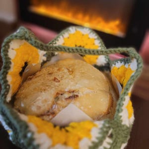 May include: A freshly baked loaf of bread in a crocheted sunflower basket. The basket is green and white with yellow sunflowers.