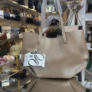 May include: A taupe-colored tote bag with dual shoulder straps, a central vertical seam, and a white product tag featuring a black abstract line drawing. The bag has a structured base and a smooth, slightly metallic finish, with protective plastic wrapping on the handles.