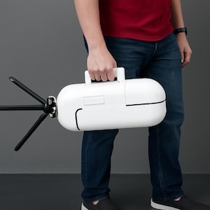 Peut inclure: Un étui de transport blanc de forme ovale avec une poignée noire et une fixation noire à plusieurs broches. L'étui est tenu par une personne portant une chemise rouge et un jean bleu. La fixation s'étend sur environ 30 cm.
