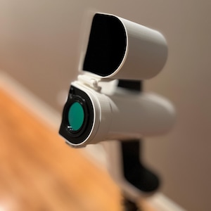 May include: A white device with a black interior and a green lens, mounted on a tripod. The device has a curved top and a cylindrical body. The background is a blurred wooden floor and a beige wall.