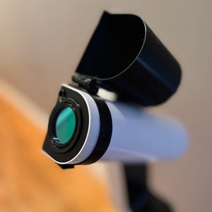 Może przedstawiać: Biały teleskop z czarną osłoną obiektywu i zielonym obiektywem. Teleskop jest skierowany w prawo.