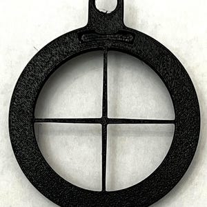 May include: A black, circular pendant with a crosshair design. The pendant has a textured surface and a loop at the top for hanging. The crosshair is formed by two intersecting lines within the circle.