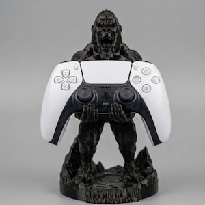 May include: A black gorilla statue holding a white and black PlayStation 5 controller. The controller has a central touchpad and button layout. The statue is detailed with a textured finish, set against a grey background.
