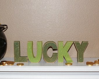 May include: Green and gold glitter letters spelling "LUCKY" on a white shelf with gold coins scattered around.