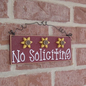 May include: A red wooden sign with white lettering that reads "No Soliciting". The sign is decorated with three yellow sunflowers with brown centers. The sign is hanging from a black wire.