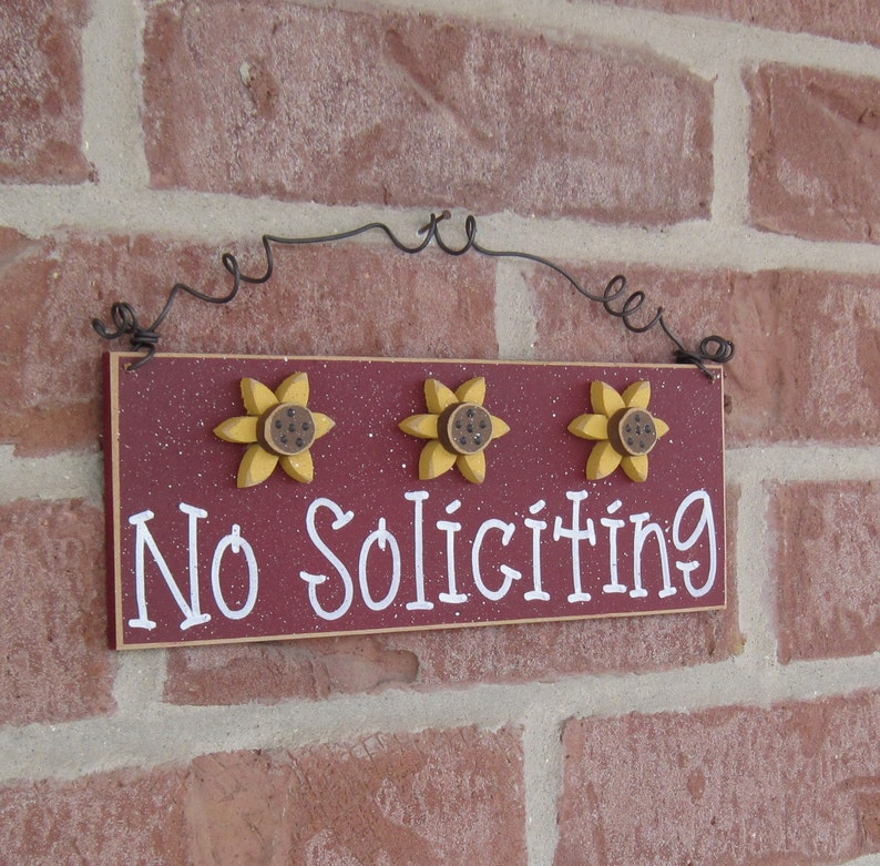 May include: A red wooden sign with white lettering that reads "No Soliciting". The sign is decorated with three yellow sunflowers with brown centers. The sign is hanging from a black wire.