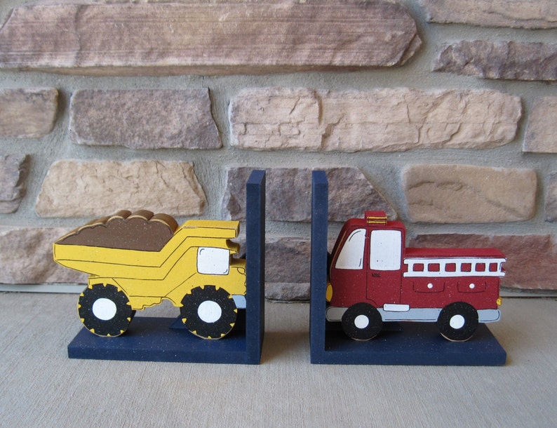 May include: A pair of wooden bookends featuring a yellow dump truck and a red fire truck. The bookends are painted with details and have a blue base.