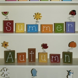 May include: Wooden blocks spelling out the words "Spring", "Summer", "Autumn", and "Winter" on a white shelf. Each word is decorated with a small, colorful, seasonal icon.