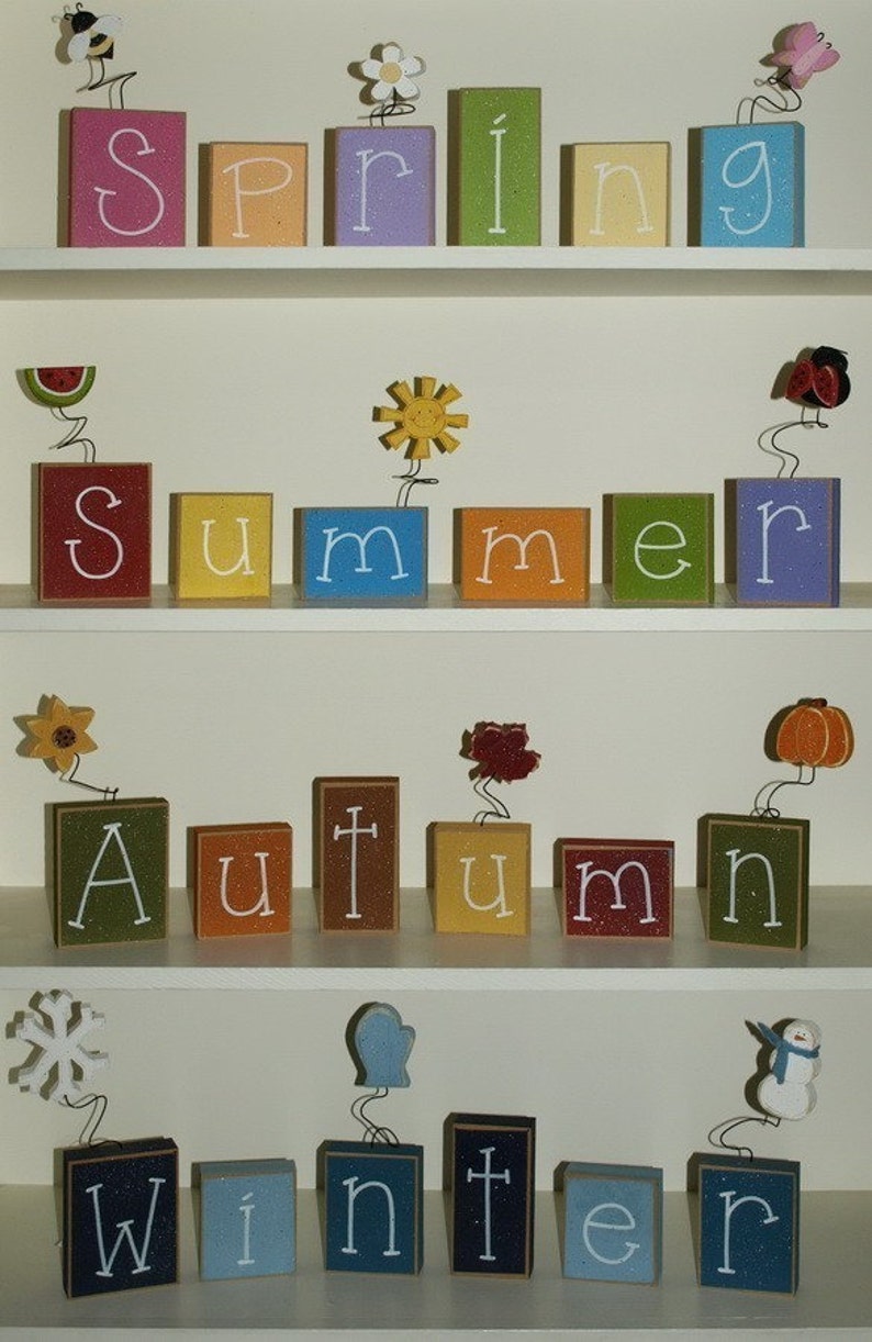 May include: Wooden blocks spelling out the words "Spring", "Summer", "Autumn", and "Winter" on a white shelf. Each word is decorated with a small, colorful, seasonal icon.