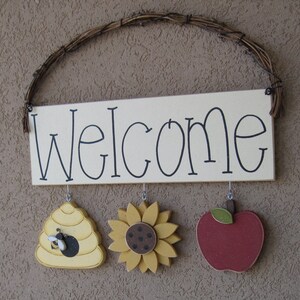 May include: A wooden welcome sign with a white background and black lettering. The sign has three hanging decorations: a yellow beehive, a yellow sunflower, and a red apple.