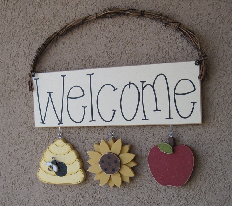 May include: A wooden welcome sign with a white background and black lettering. The sign has three hanging decorations: a yellow beehive, a yellow sunflower, and a red apple.