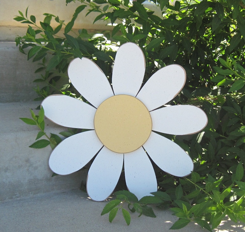 May include: A large white daisy with a yellow centre, made of wood, is placed on a concrete step. The daisy is surrounded by green leaves.